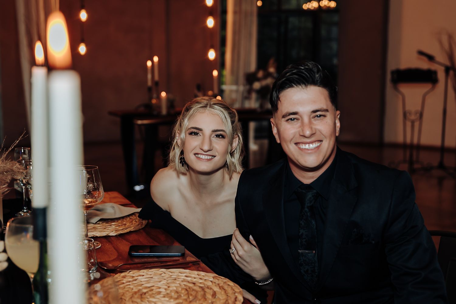 A couple smiles together at an elegant candlelit dinner table with warm ambient lighting in the background.
