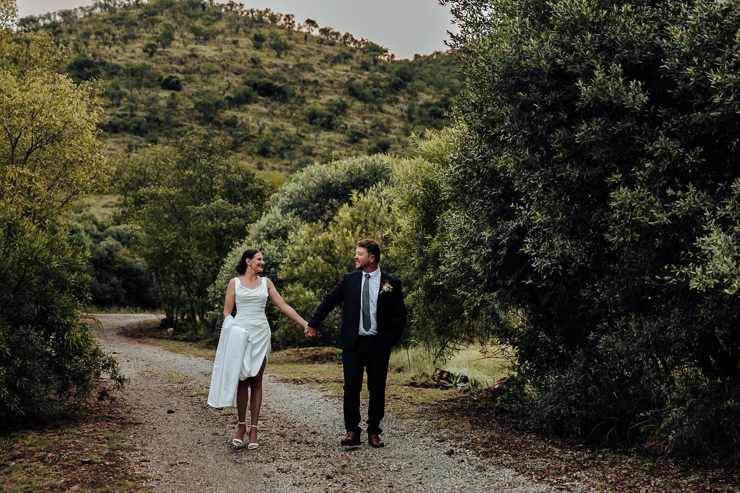 A couple walks hand in hand down a scenic path surrounded by green foliage and natural landscape.