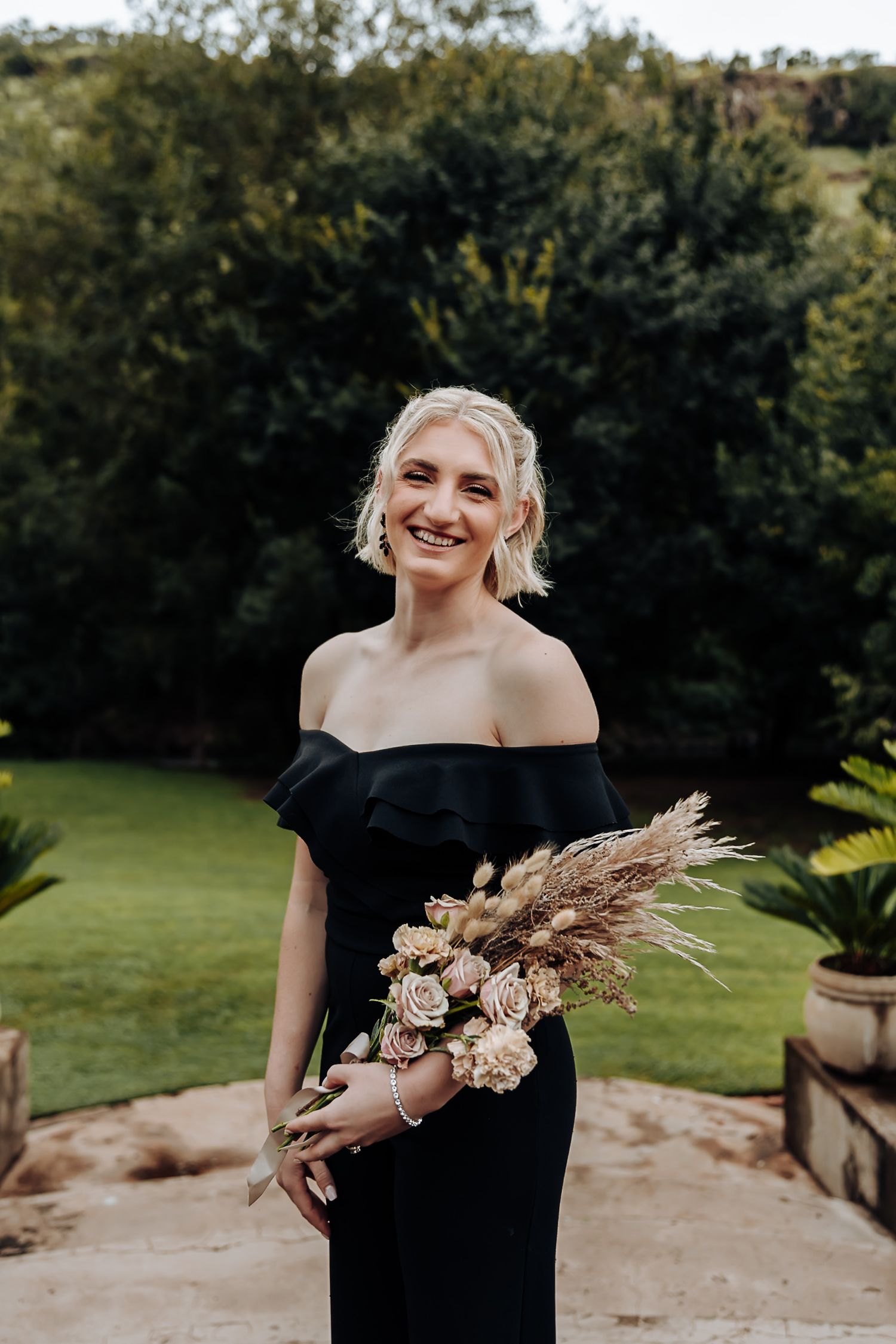 Someone in an elegant black off-shoulder dress holds a dried floral bouquet while standing in a garden setting.