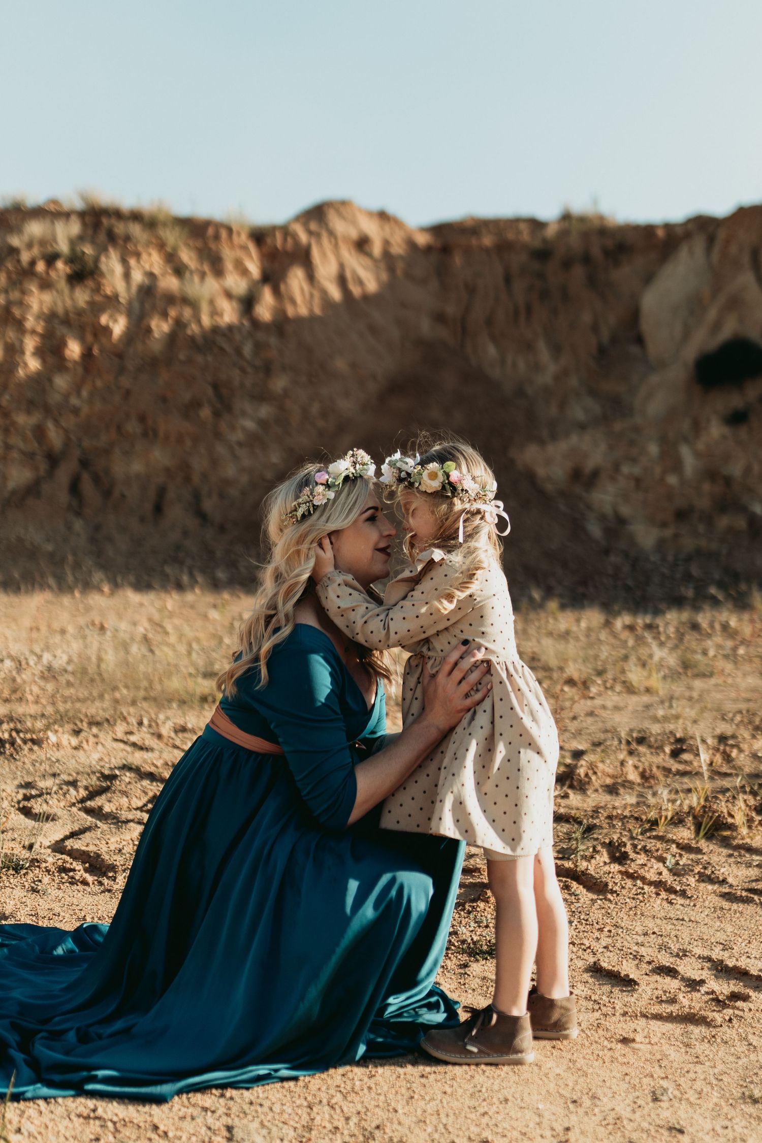 Person in blue dress sits on desert ground interacting lovingly with a gentle alpaca.