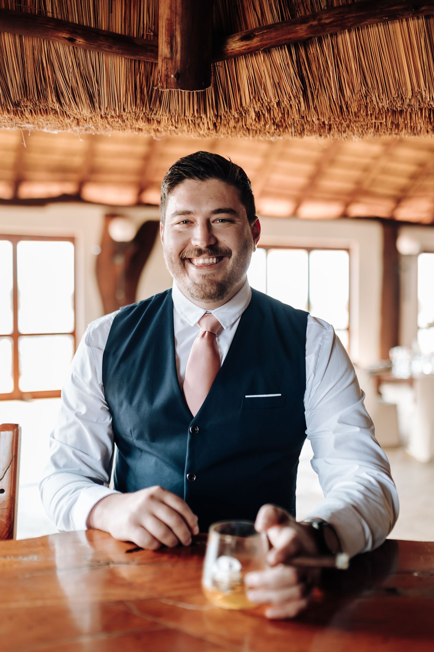 Man in business attire smiles while sitting at wooden bar in restaurant setting with warm lighting.