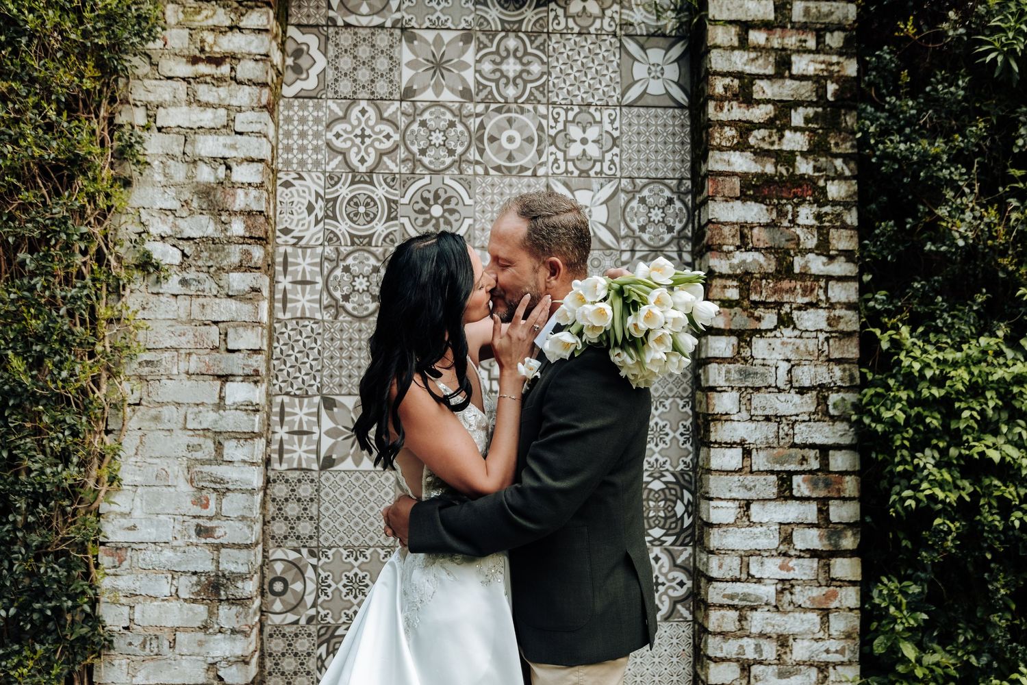 Bride and groom share a romantic kiss in front of an ornate tiled wall surrounded by lush greenery.