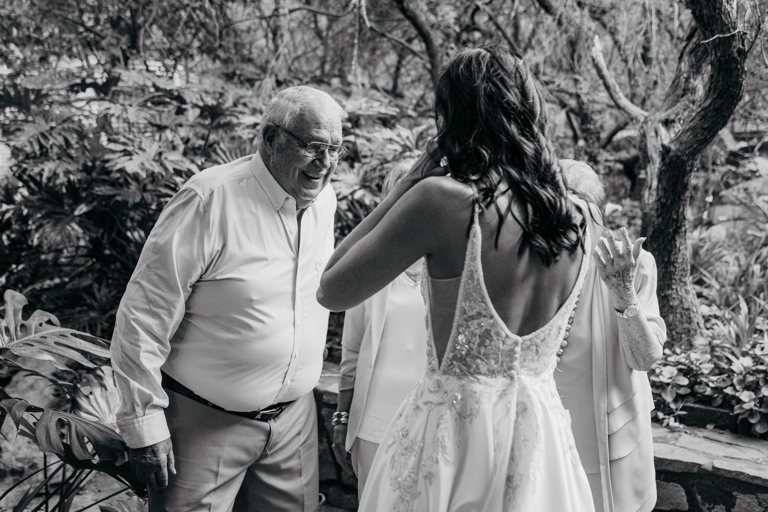 Black and white photo of a bride in lace gown conversing with an older man outdoors amid lush greenery.