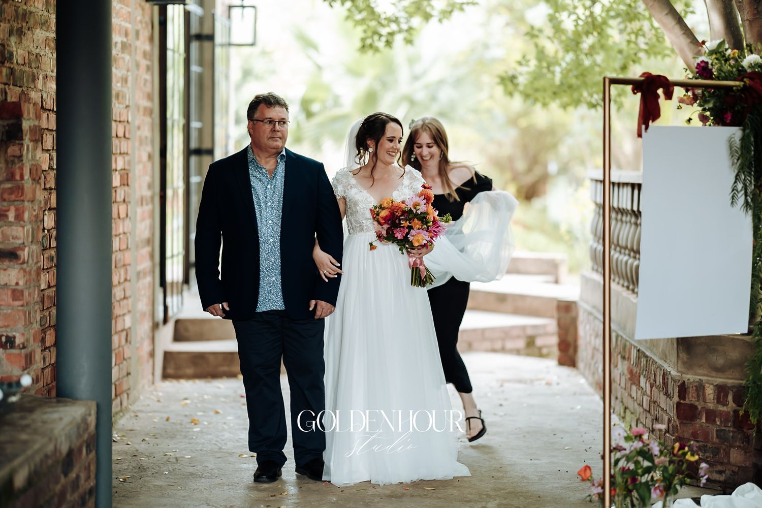 Bride in white gown walking with two escorts along a covered outdoor walkway.