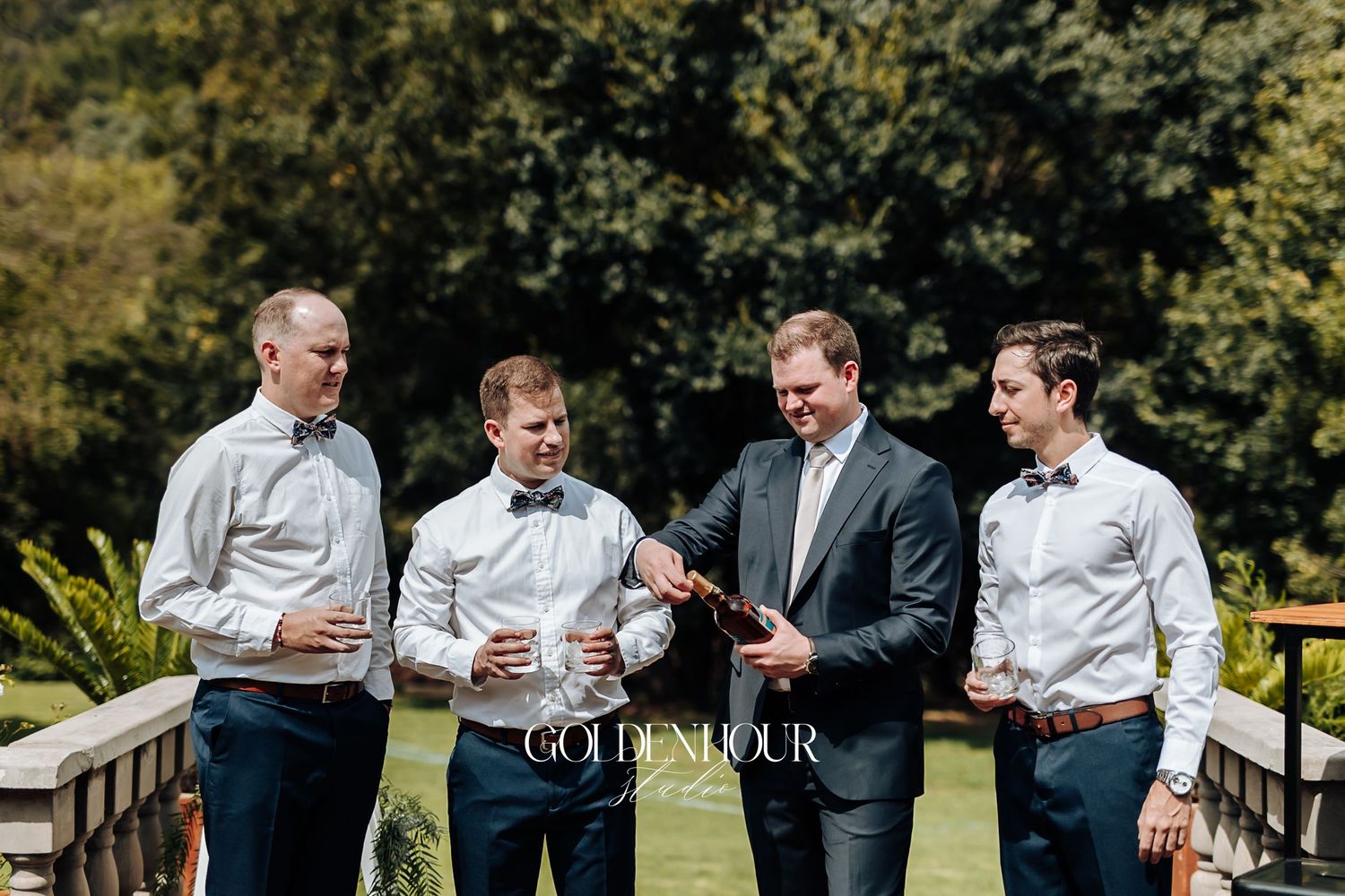 Groom and groomsmen in white shirts and navy pants celebrating outdoors with champagne before the wedding.