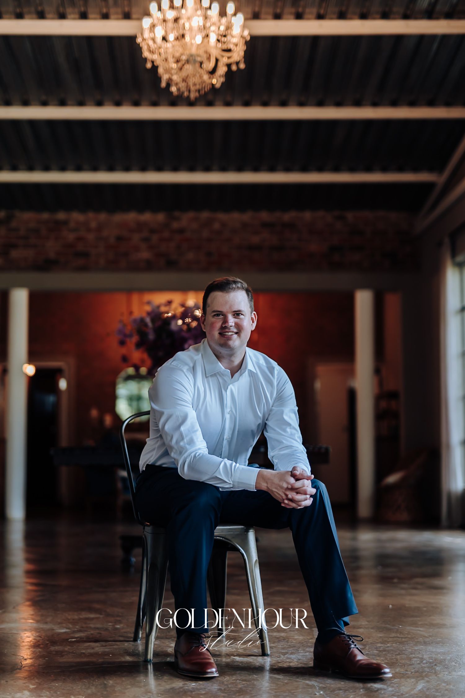 Groom sits smiling on a metal chair inside an elegant venue with chandelier and brick walls.