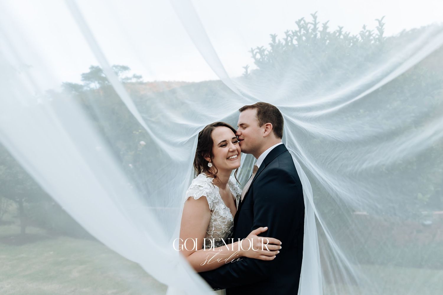 Bride and groom embrace romantically under a flowing white veil outdoors.