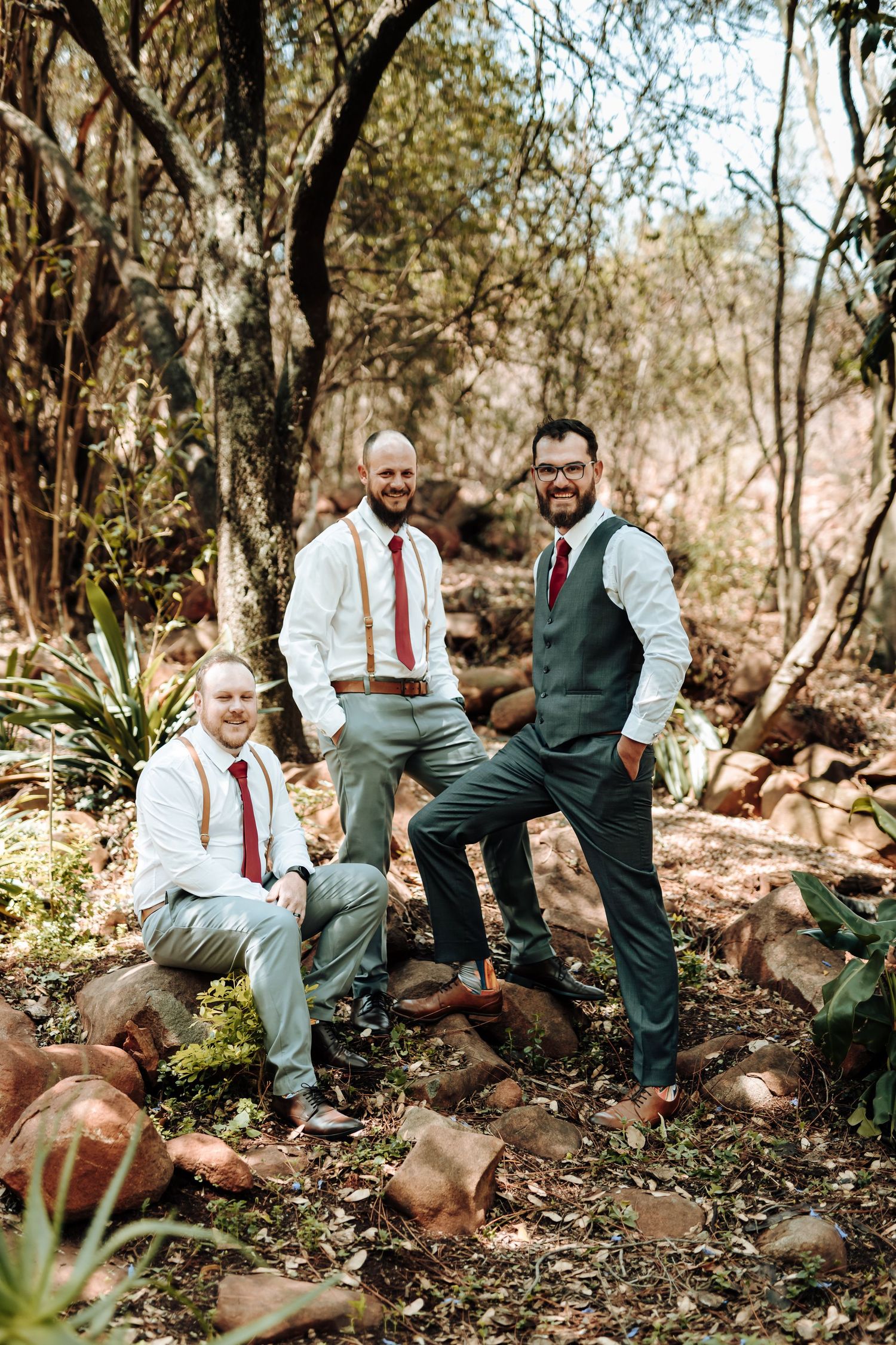 Three groomsmen in matching outfits pose on natural rock formation in outdoor setting.