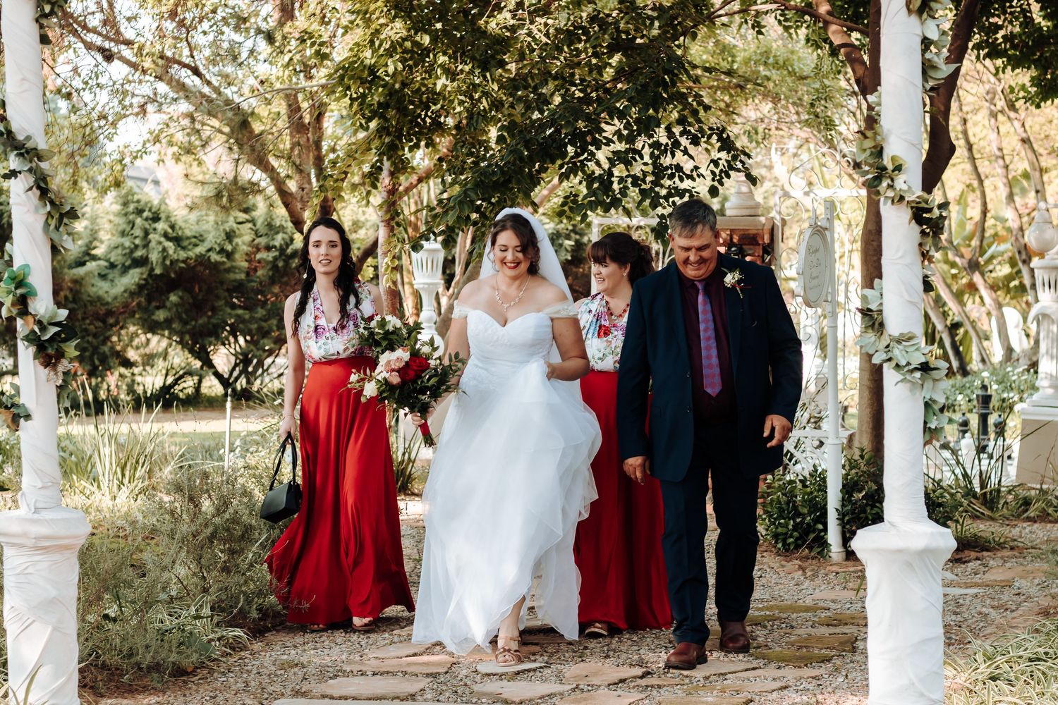 Outdoor wedding processional under white pillars with bridesmaids in red dresses.
