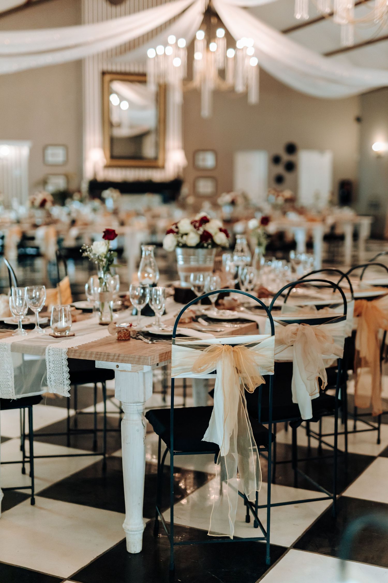 Wedding venue with black and white checkered floor, white chair sashes, and crystal chandeliers.