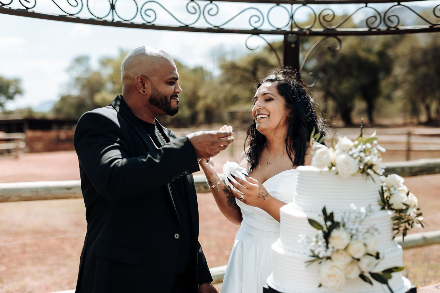Wedding couple laughing while feeding each other cake in an outdoor setting with white floral decorations.