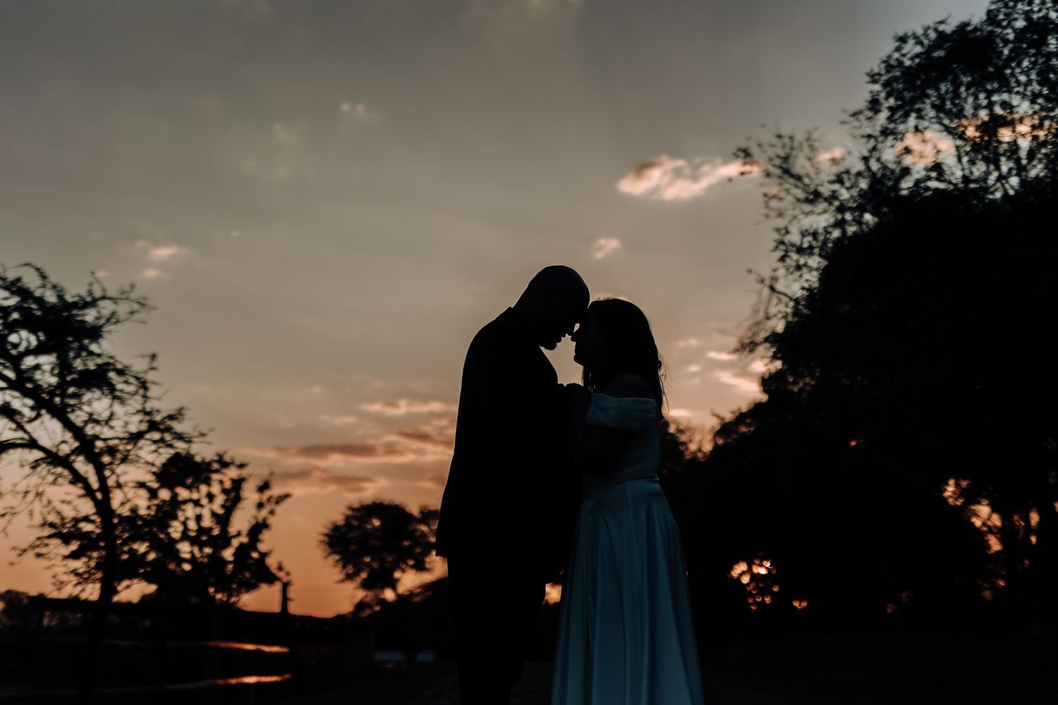 Silhouetted couple shares intimate moment against dramatic sunset sky during wedding.