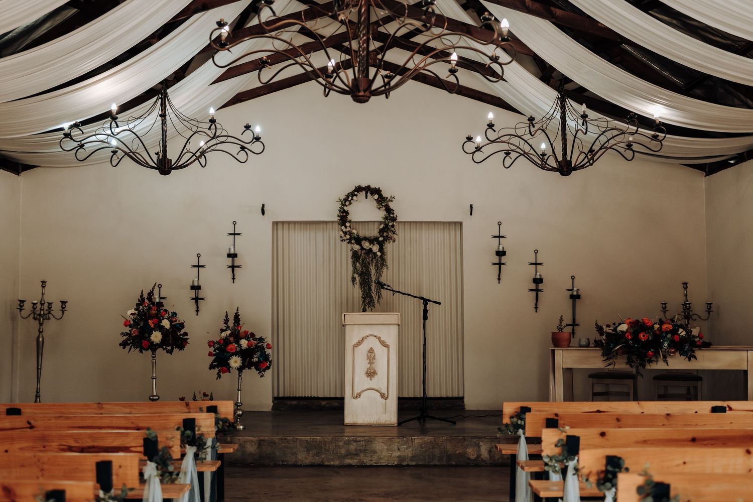 Interior of a rustic chapel with wooden pews, draped fabric ceiling, and elegant chandeliers for a wedding ceremony.