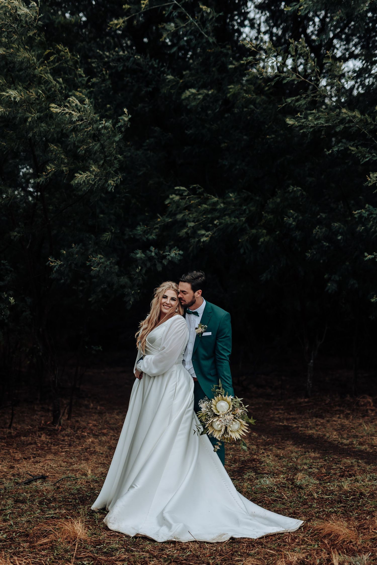Moody forest wedding photo with couple embracing, bride holding white and green bouquet.