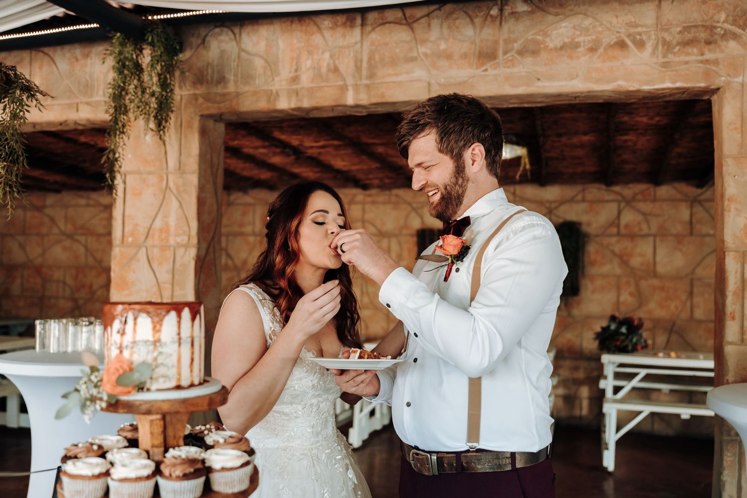 Couple playfully feeds each other cupcakes at their wedding reception under a rustic wooden beam.