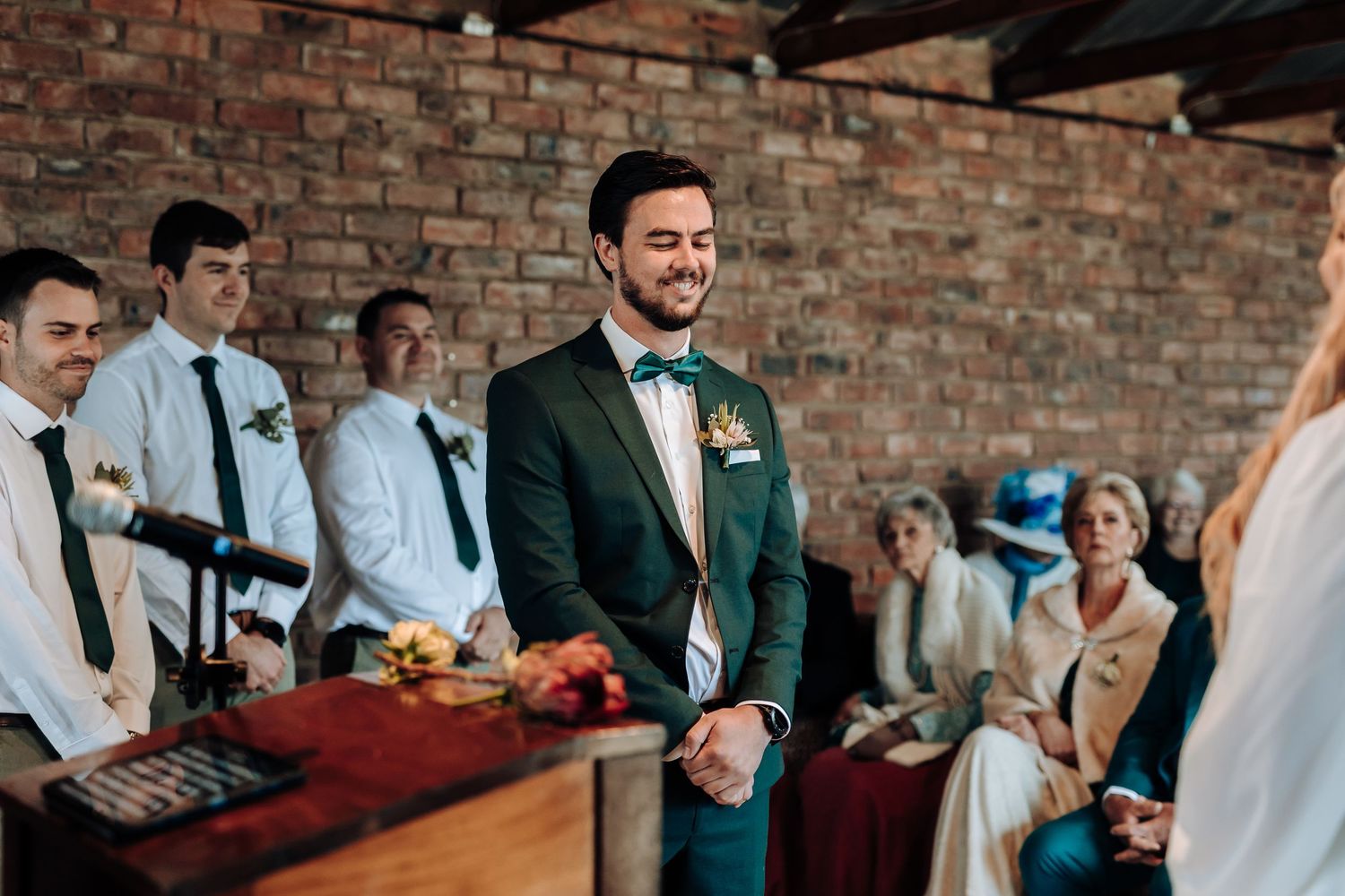 A groom in emerald green tuxedo stands at altar during wedding ceremony in rustic brick venue.