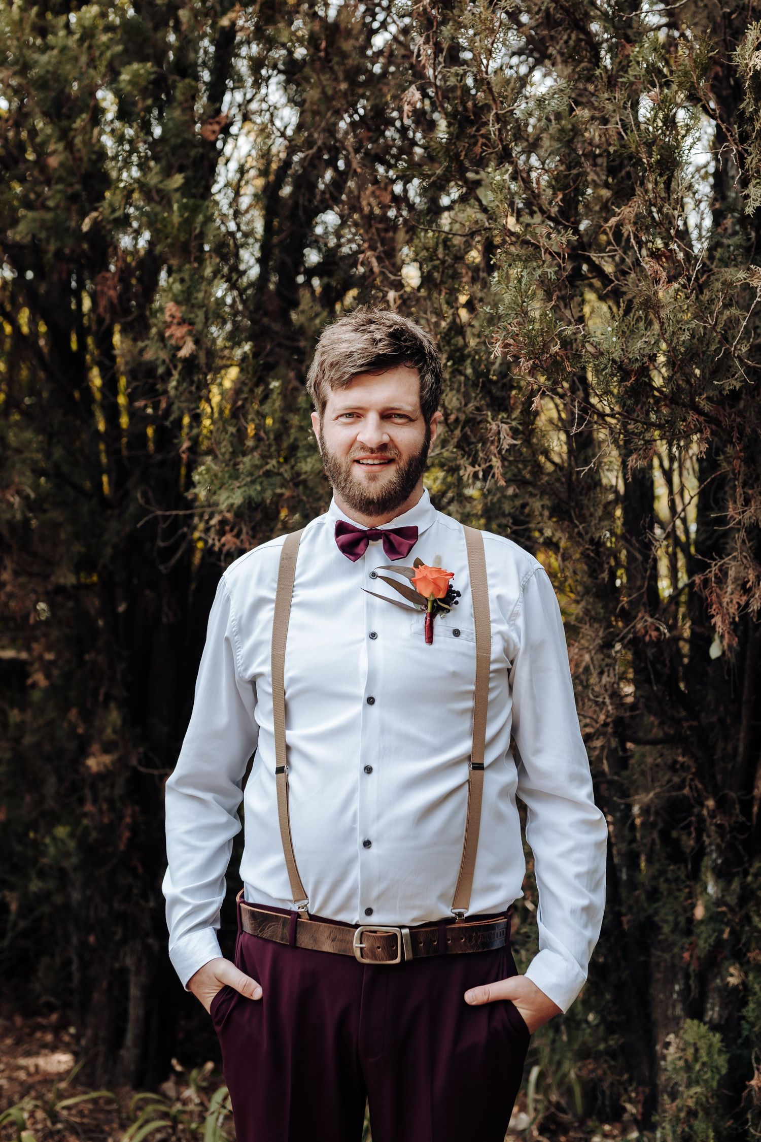 Groomsman wearing burgundy pants and brown suspenders poses in front of trees.