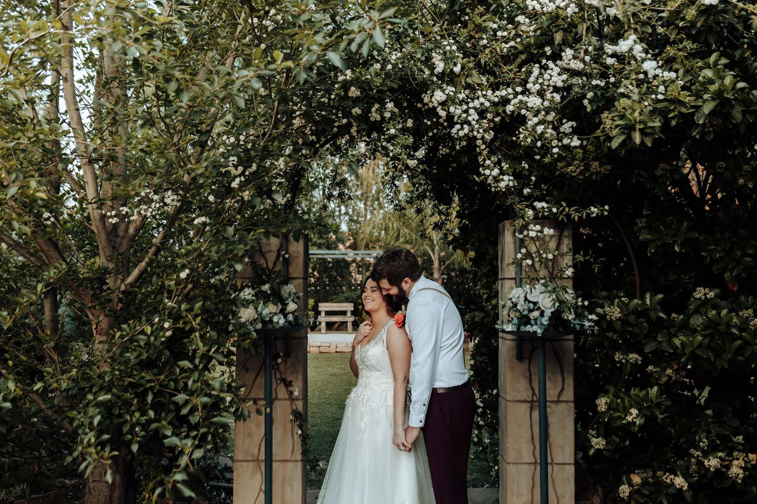 Couple embracing under floral archway in garden wedding setting.