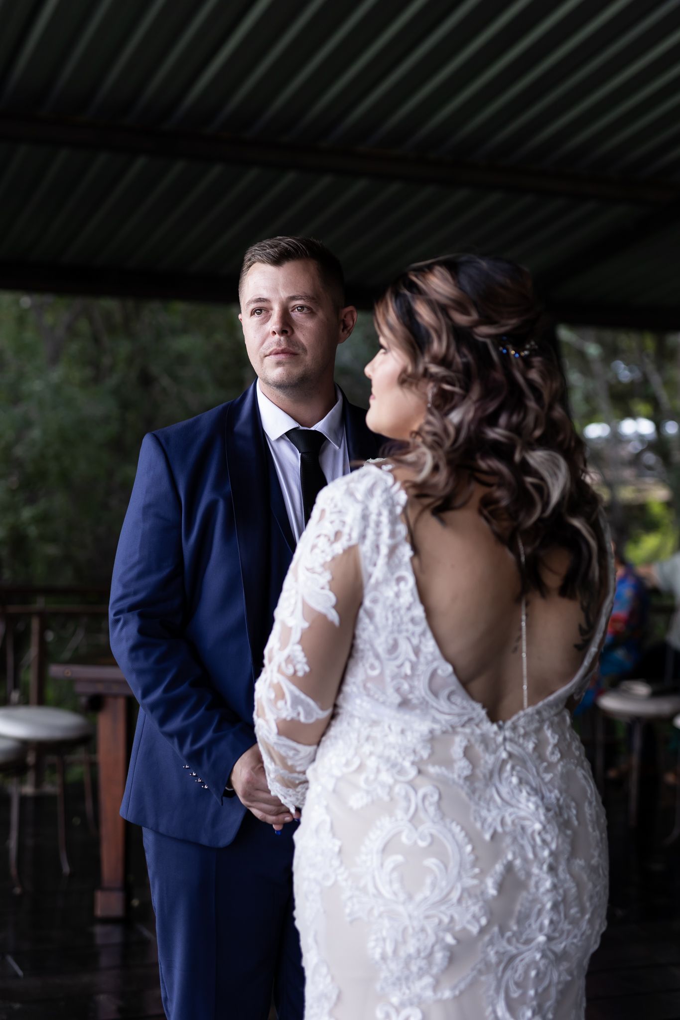 Elegant bride in lace gown with open back stands with groom in navy suit.