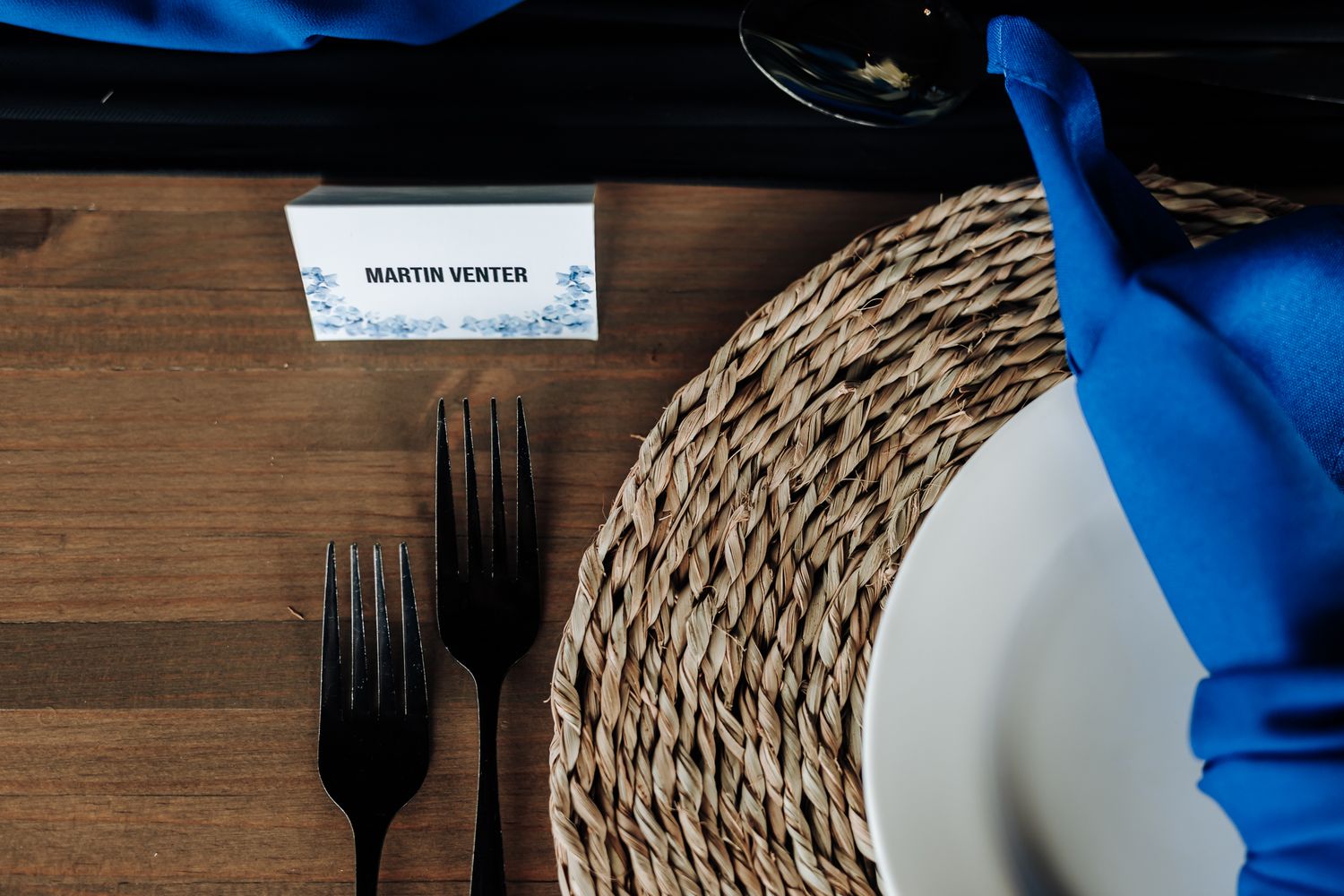 Place setting with blue napkin, woven placemat, and black forks on wooden table.