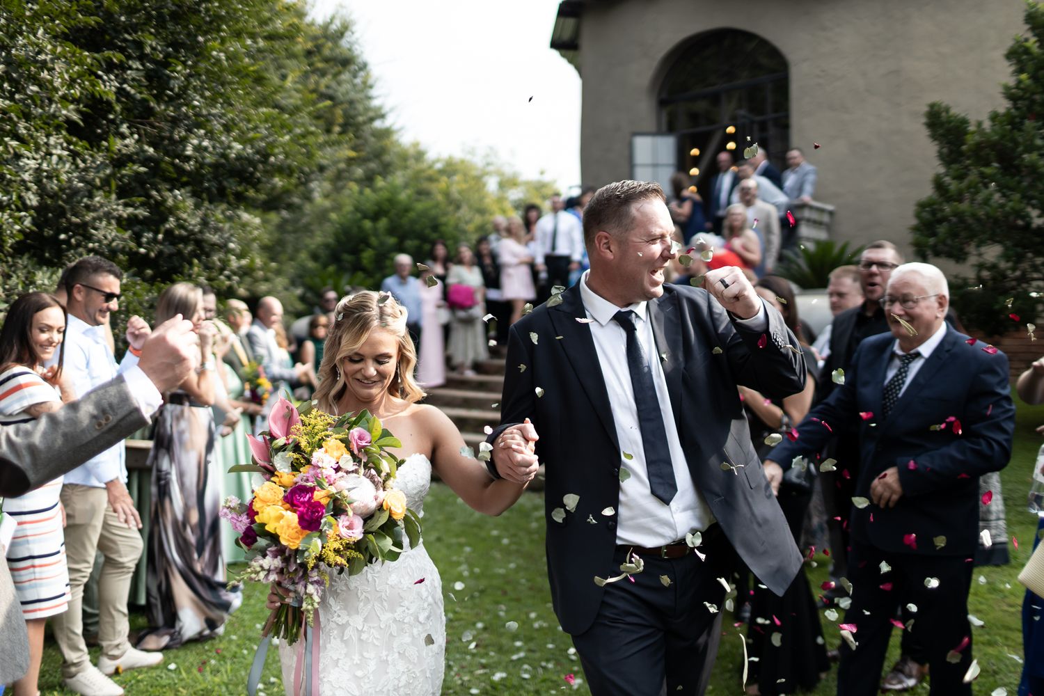 Joyful couple celebrates while walking through crowd of guests throwing confetti outside church.