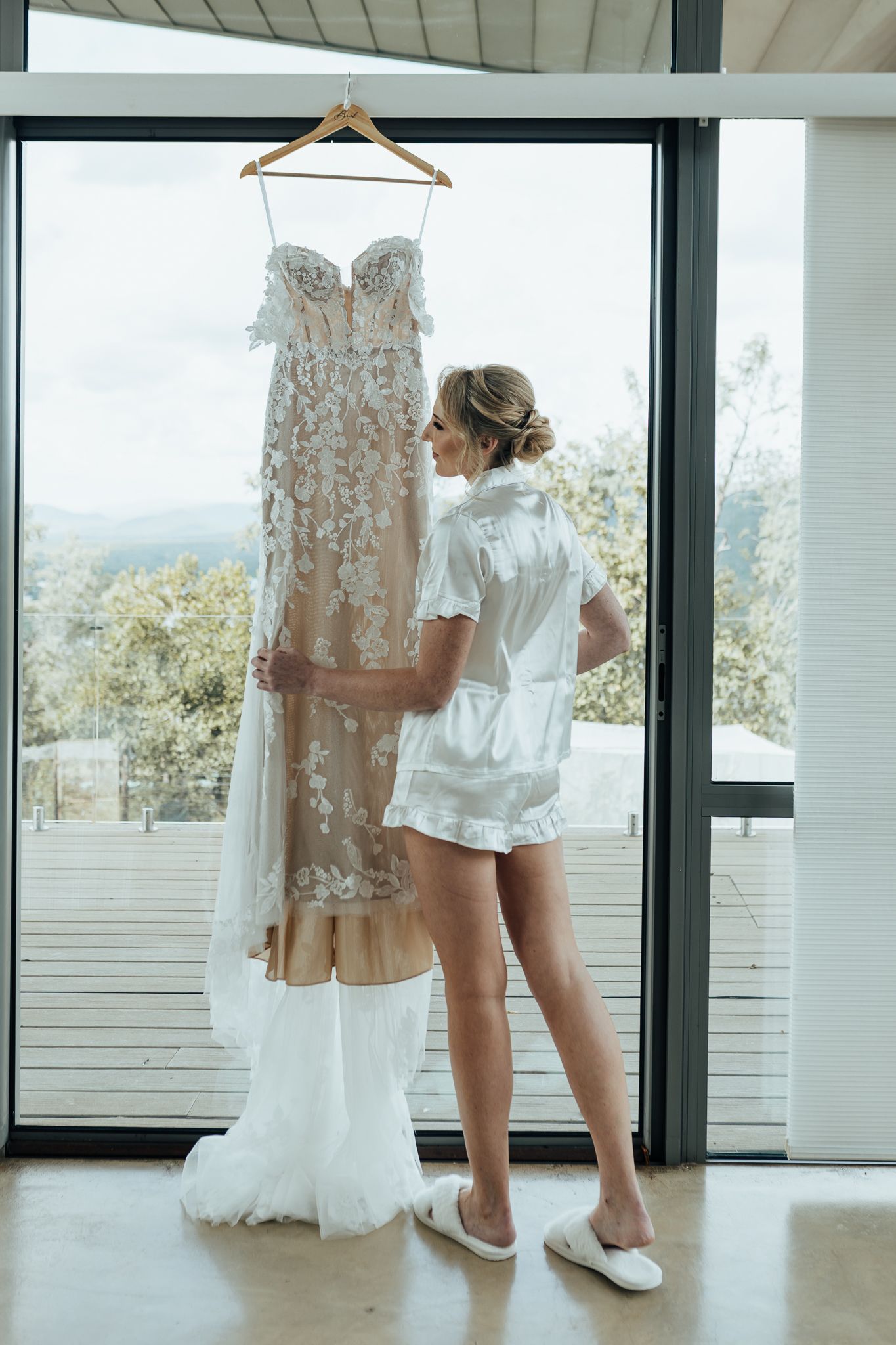 Wedding dress hanging by window while someone in white robe admires intricate lace details.