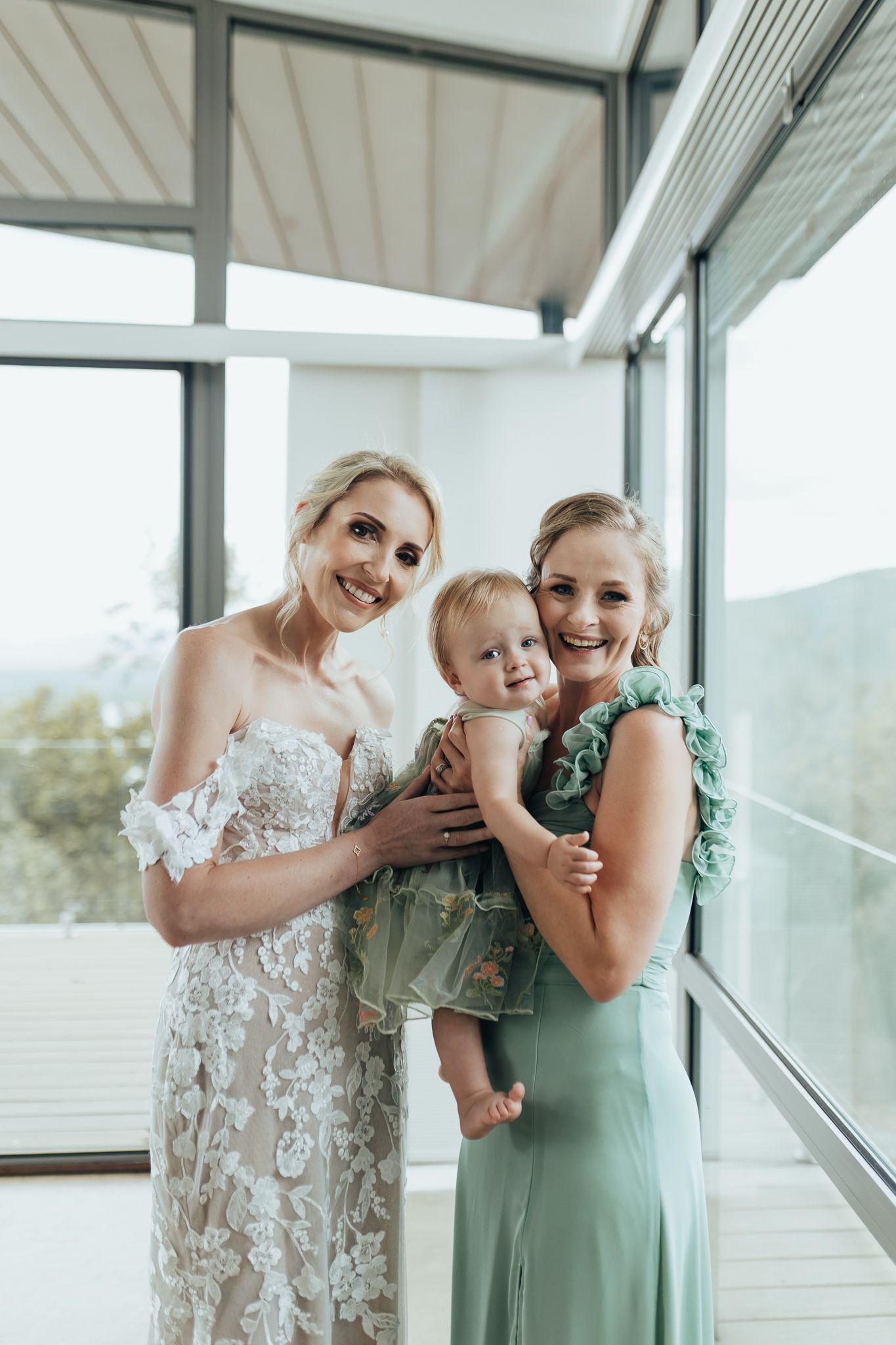 Wedding party stands in sunlit room with mountain views through large windows.