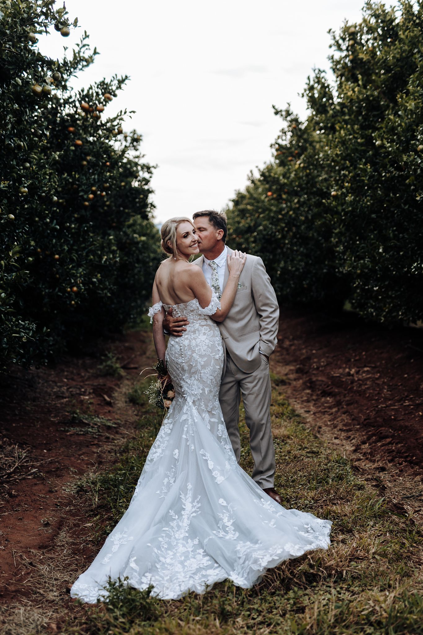 Elegant wedding couple embraces in a citrus orchard with stunning lace dress details.