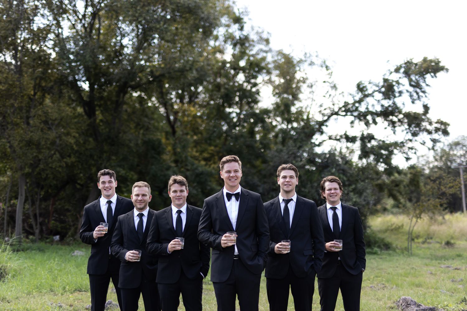 Group of groomsmen in black suits posing together in outdoor setting.