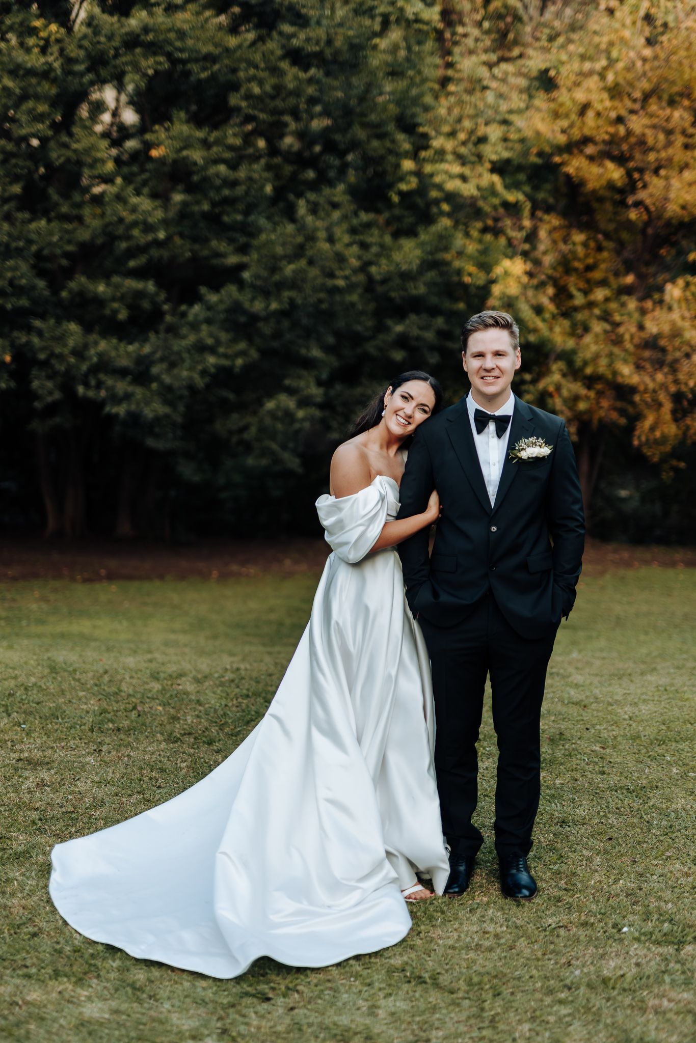 Elegant wedding couple poses in formal attire against autumn landscape.