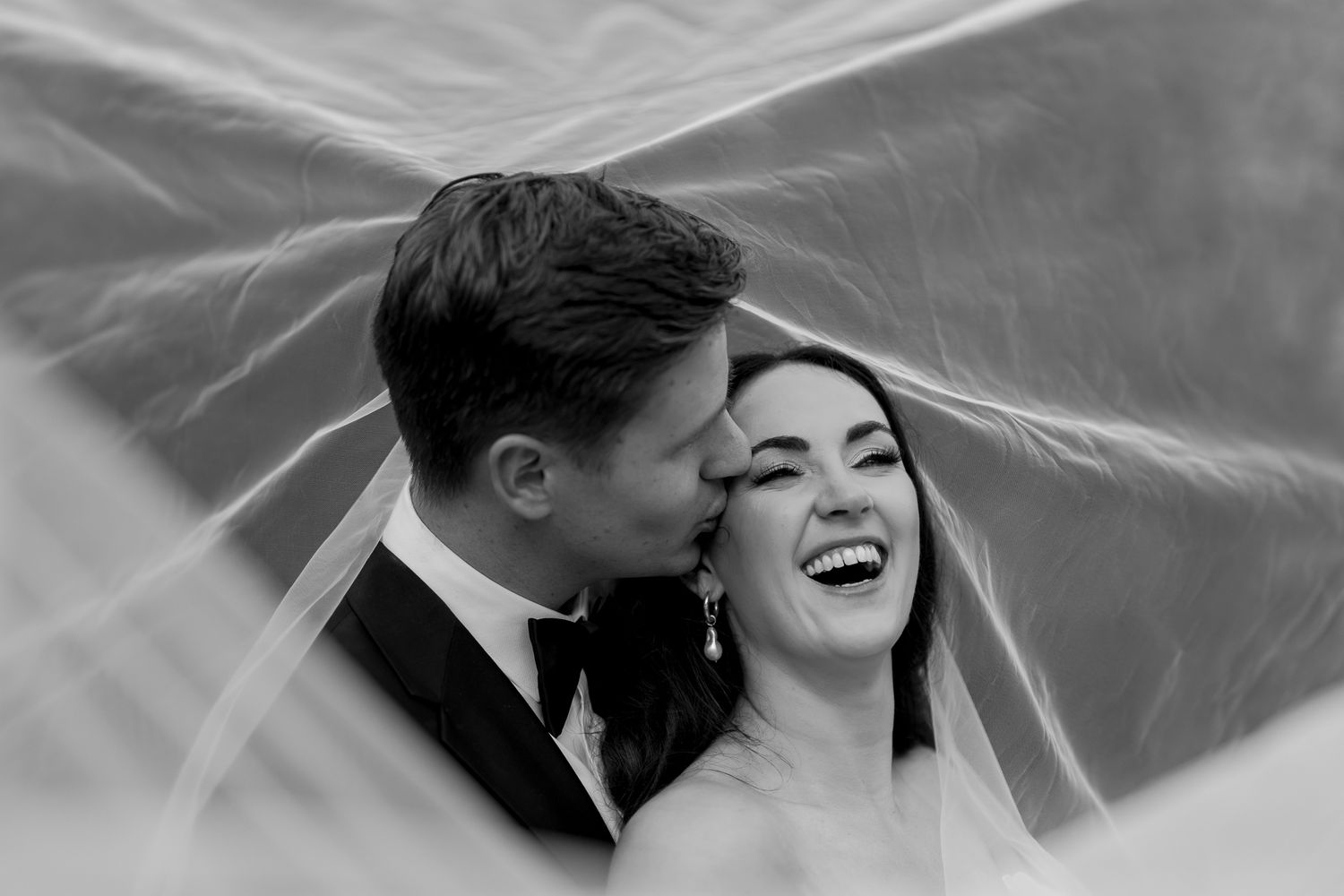 A joyful black and white moment captured under flowing wedding veil fabric.