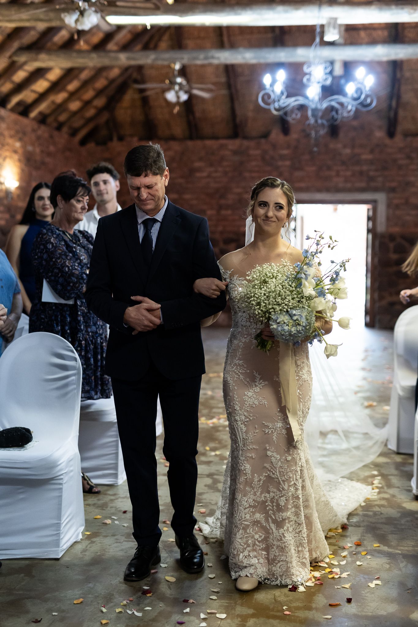 Bride in lace gown walks down aisle with escort in rustic brick venue with chandelier overhead.