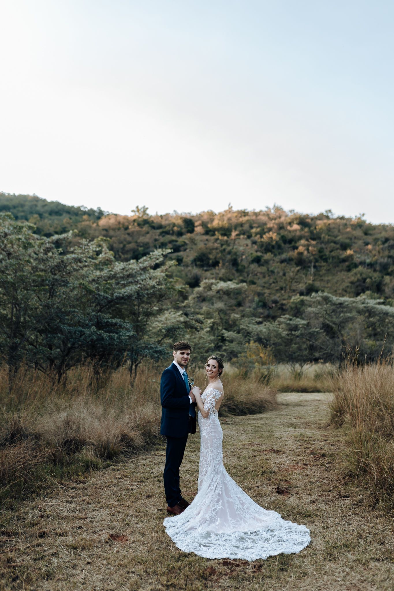 A wedding couple stands together on a dirt path with a dramatic hillside landscape behind them.