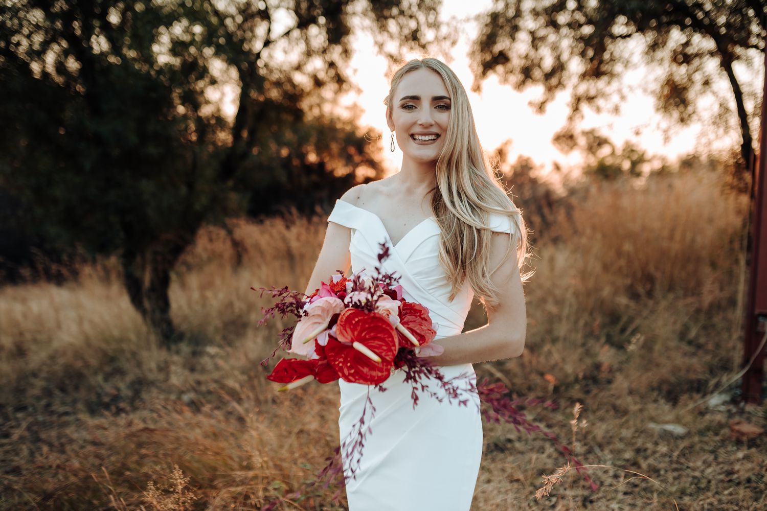 A bride in a fitted white dress holds a red bouquet against a sunset landscape.