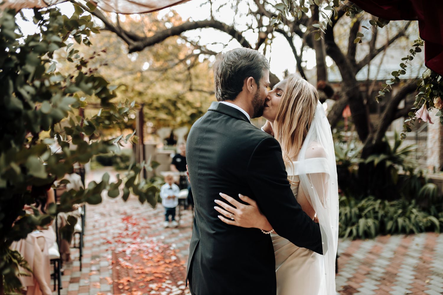 Romantic wedding couple embracing under autumn trees with rose petals scattered on brick pathway.