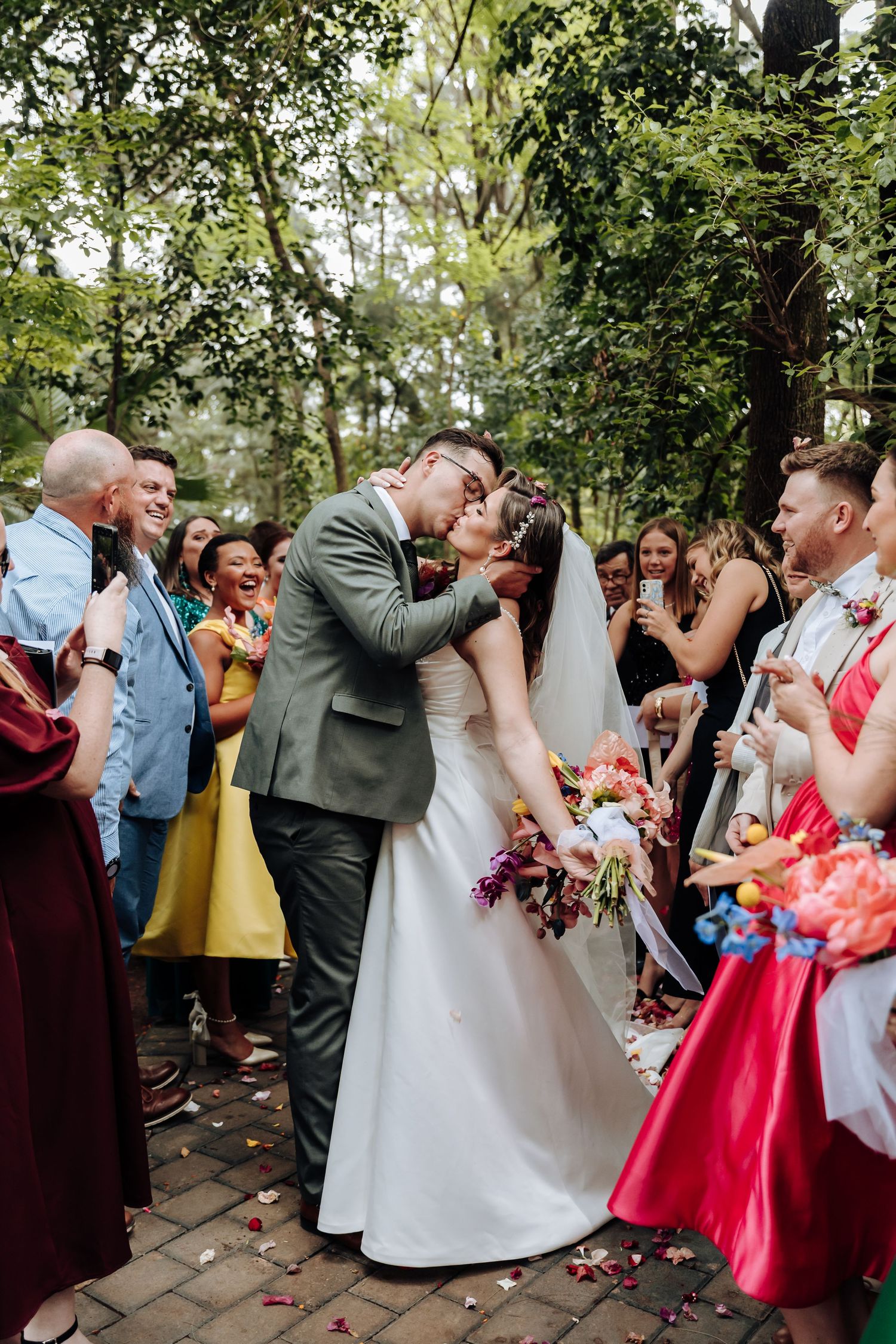 Newlyweds share their first kiss surrounded by cheering guests on a tree-lined outdoor pathway.