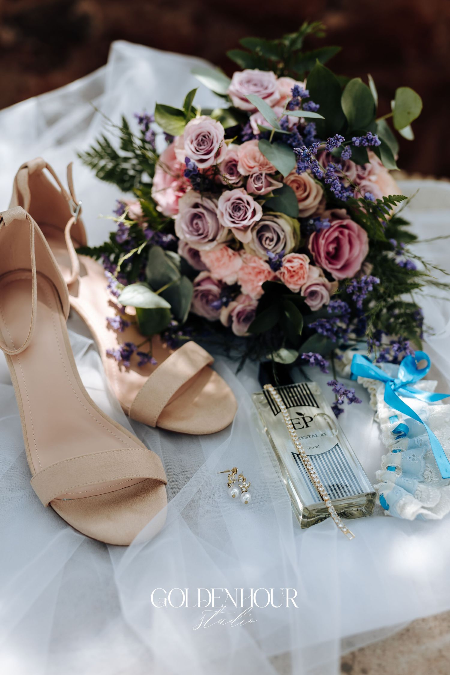 Wedding details including blush roses, blue flowers, and nude bridal shoes arranged on white fabric.