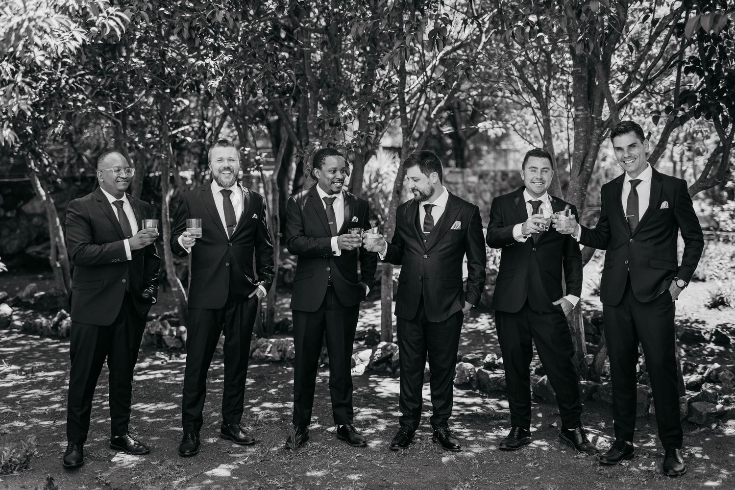 Group of groomsmen in black suits posing together outdoors in a natural wooded setting.