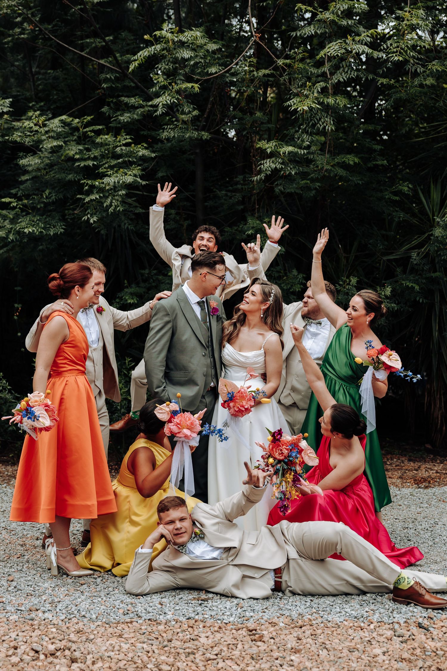 Wedding party in colorful dresses poses playfully together on a garden path with trees in background.
