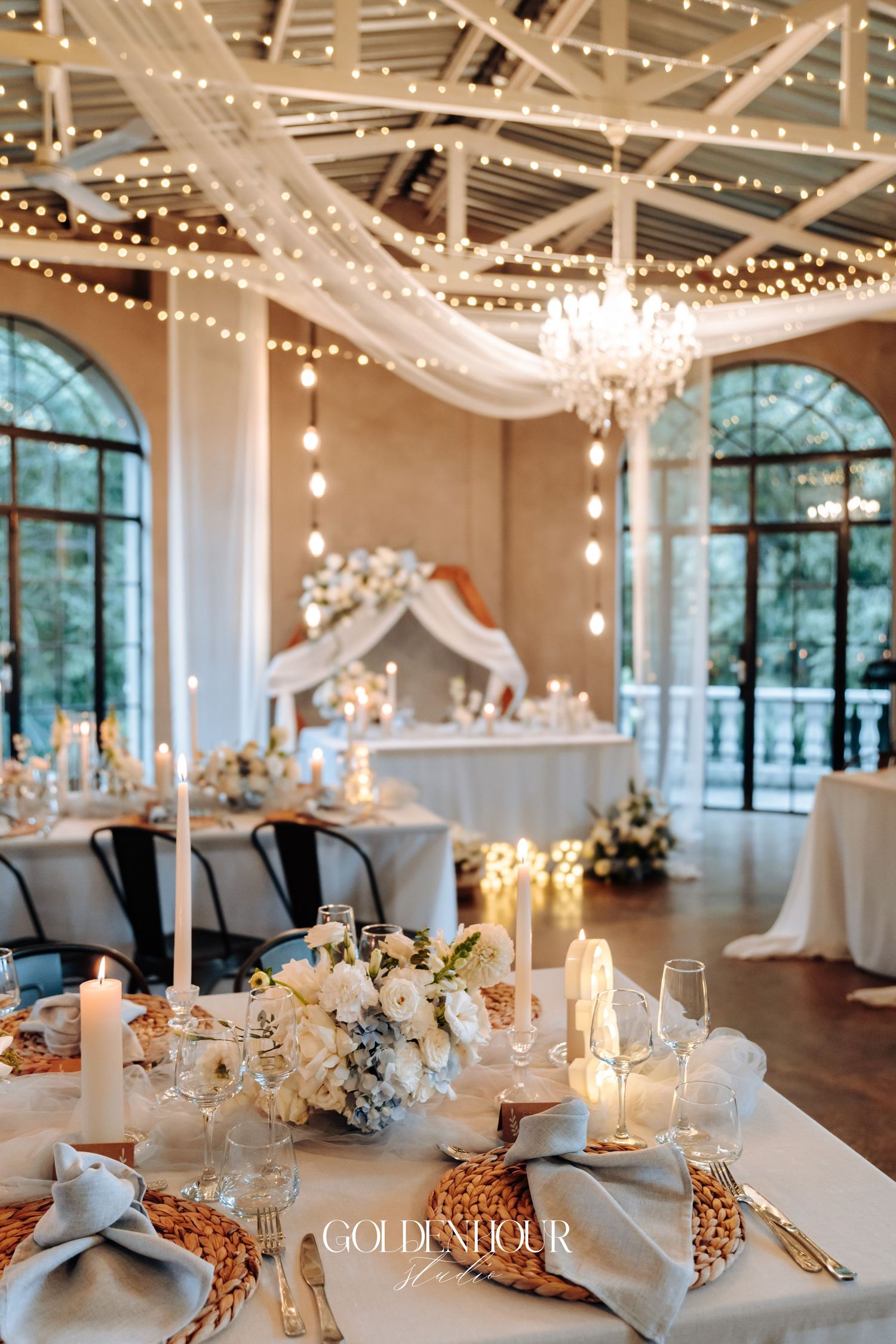 Elegant wedding reception venue with string lights, white linens, and arched windows creating a romantic atmosphere.