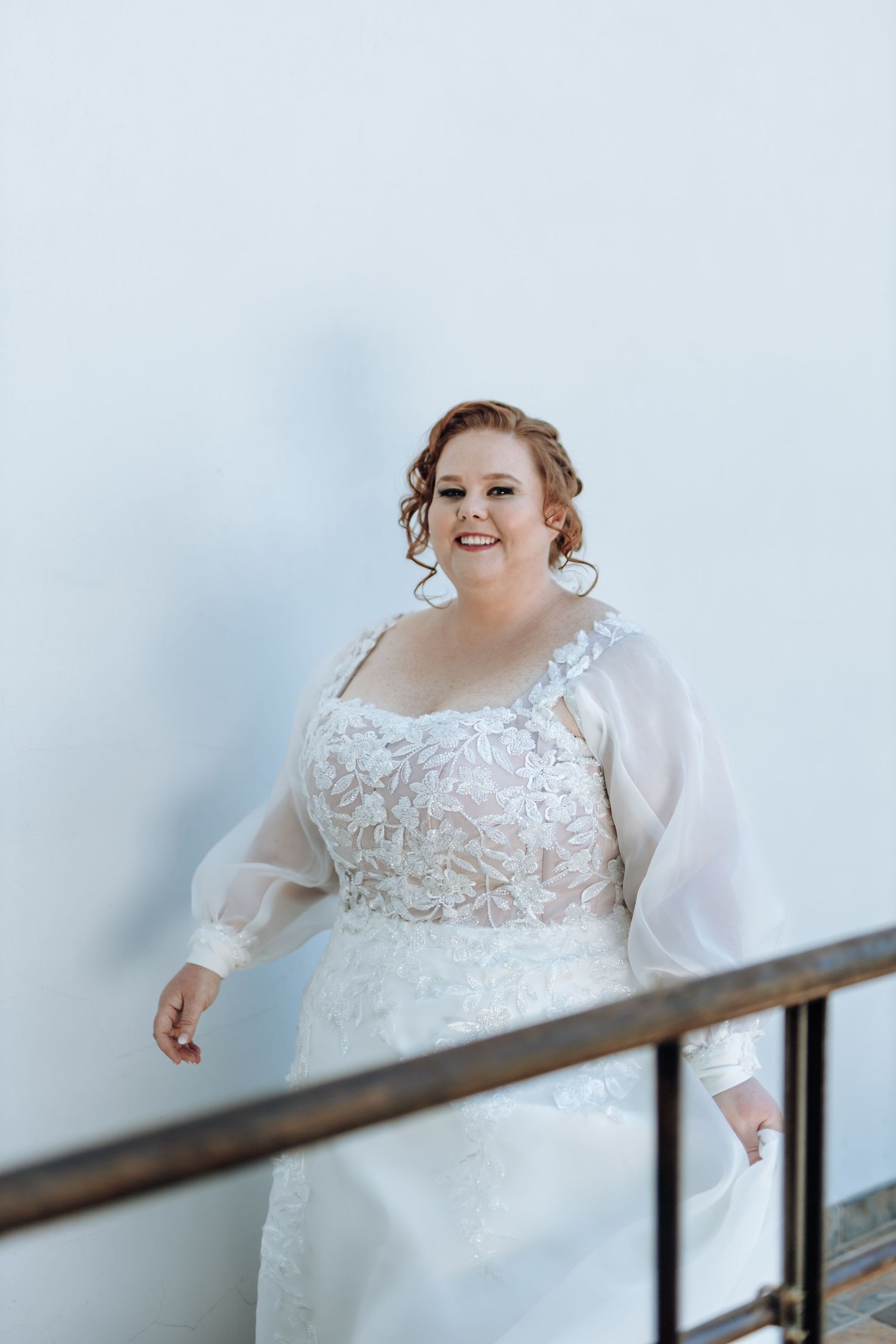 A person in a white lace wedding dress with sheer sleeves stands smiling by a railing against a light background.