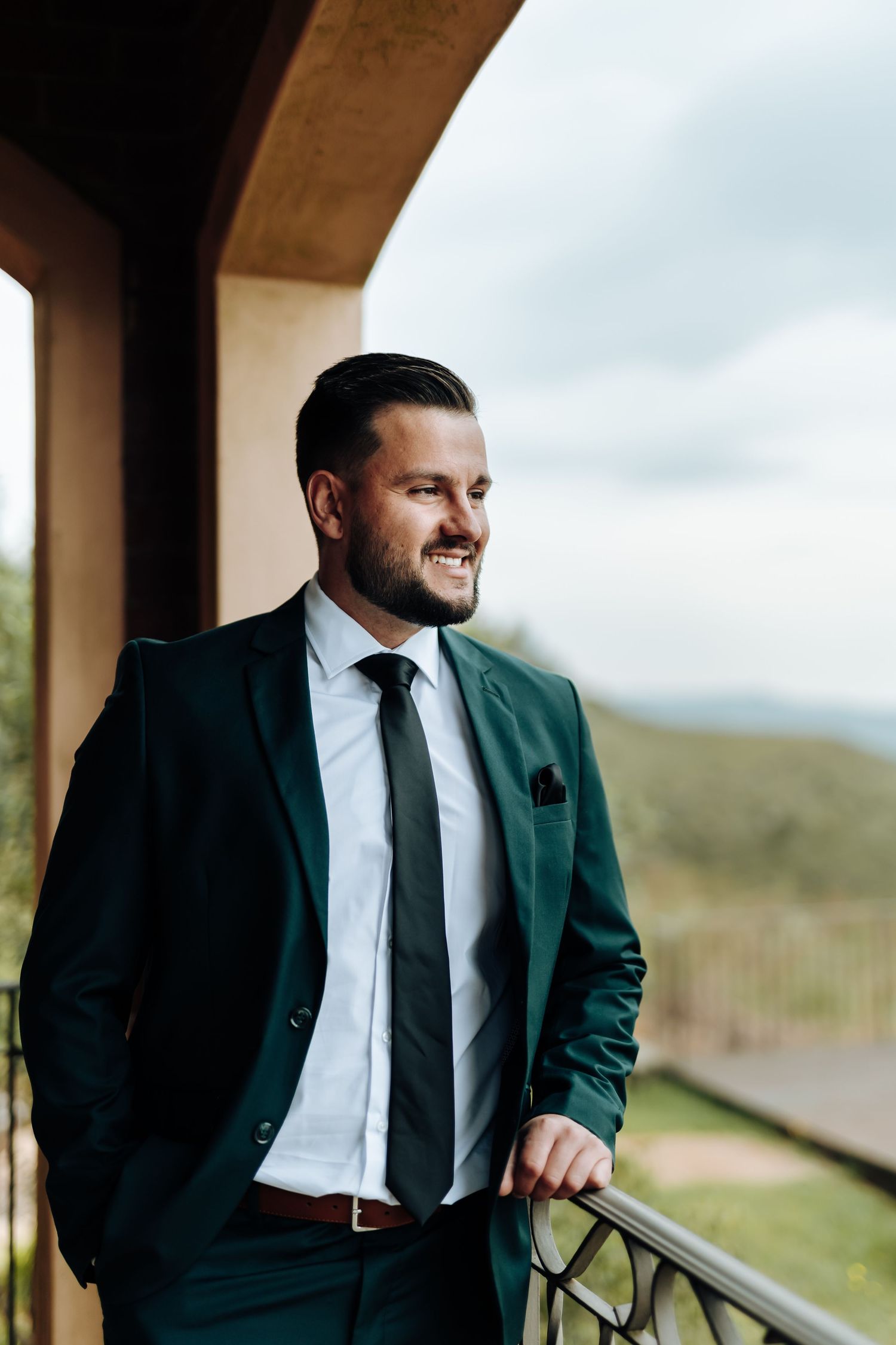Groom in dark green suit jacket and black tie smiles while looking out from covered porch on cloudy day.