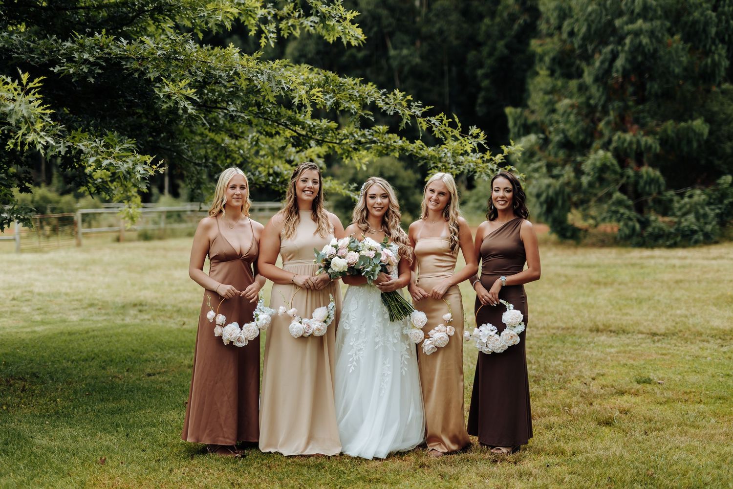 Wedding party in brown and beige dresses poses together on a sunny grassy lawn during outdoor celebration.