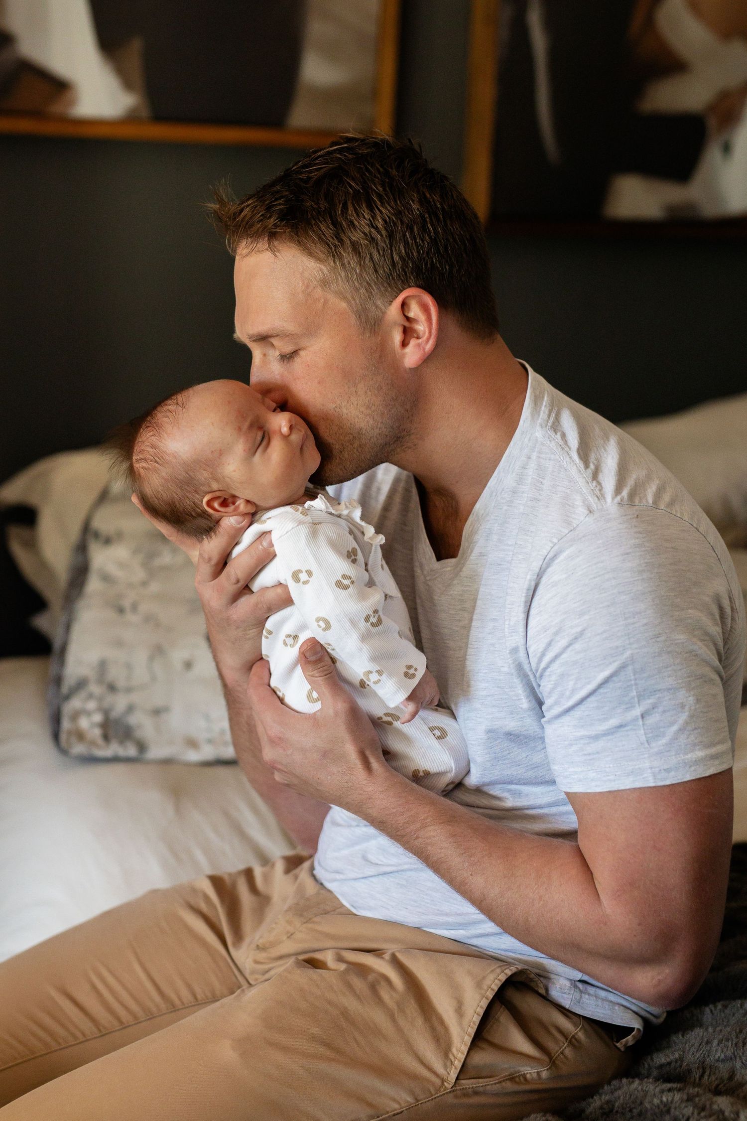 A tender moment shows a parent in a grey t-shirt gently kissing a newborn baby wearing white onesie while sitting on a bed.