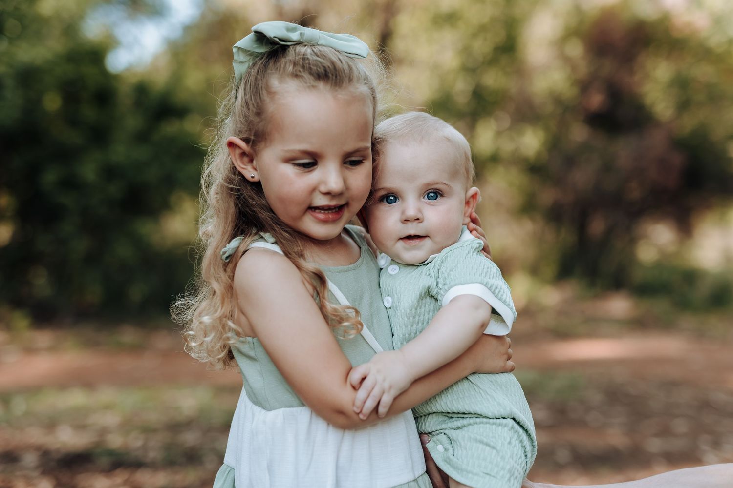 Two young children share a loving embrace while posing outdoors in light colored clothing.