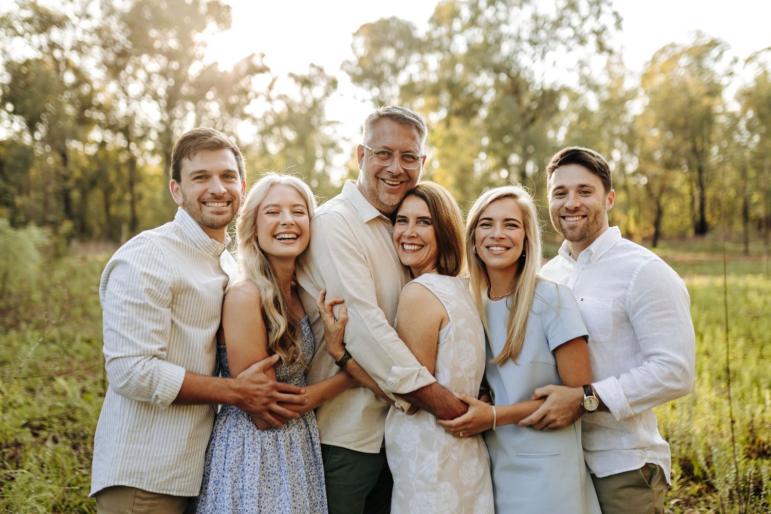 Extended family shares laughter and embraces while wearing coordinated white and beige outfits in a natural outdoor setting.