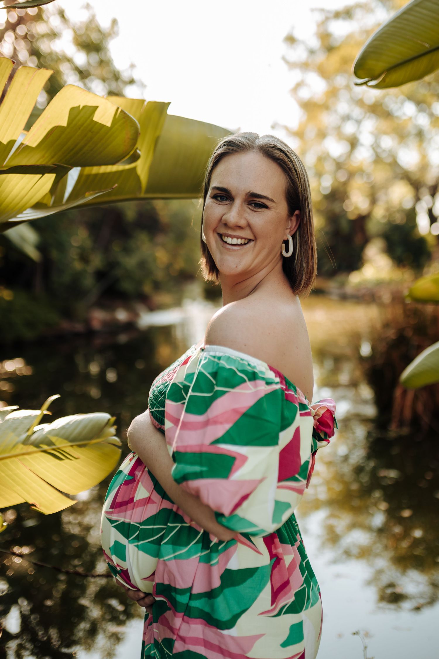 A smiling figure poses in a vibrant tropical print dress by a pond with banana leaves framing the scene.