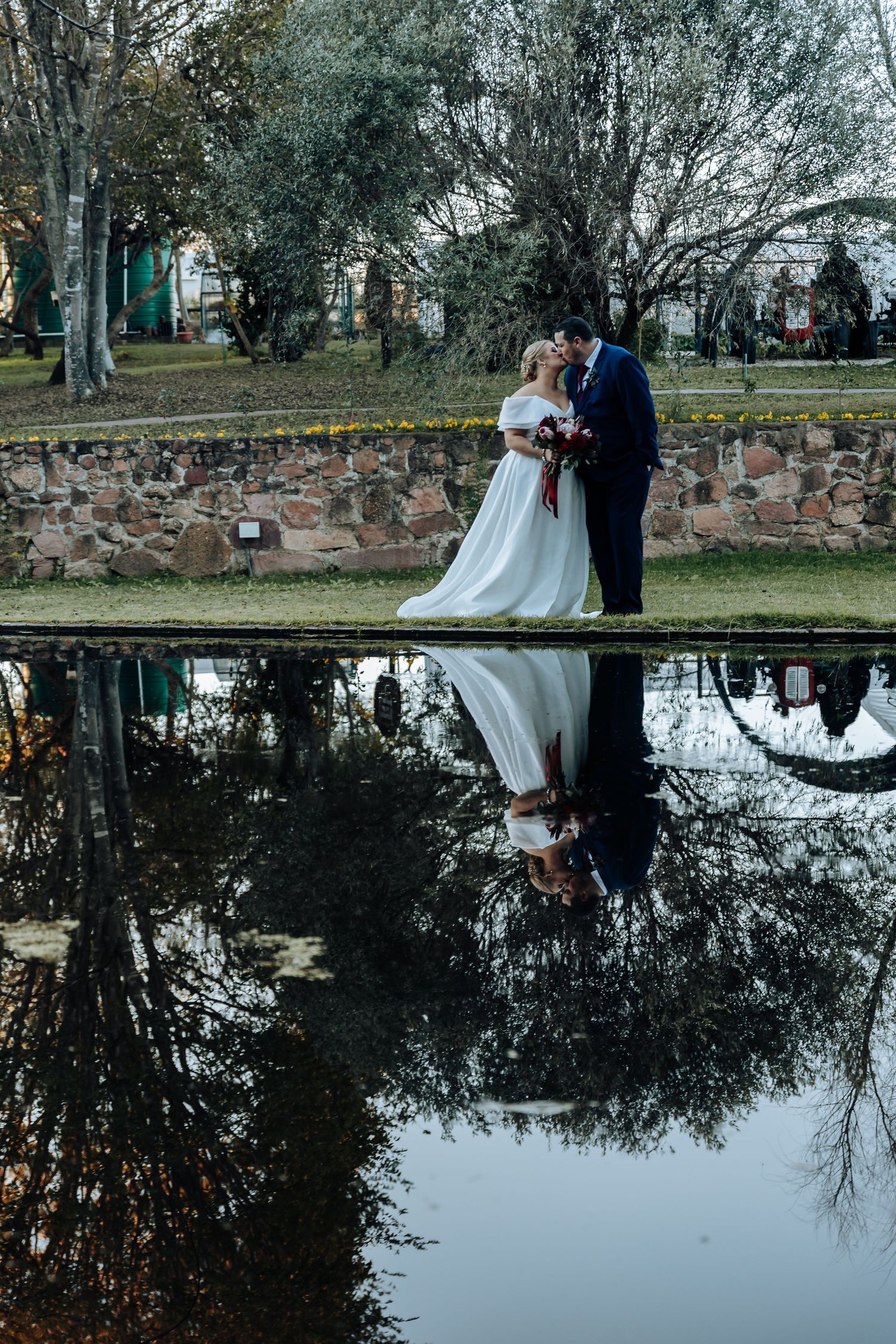 Bride and groom kissing by a reflective pond in a park, their reflection mirrored in the still water.