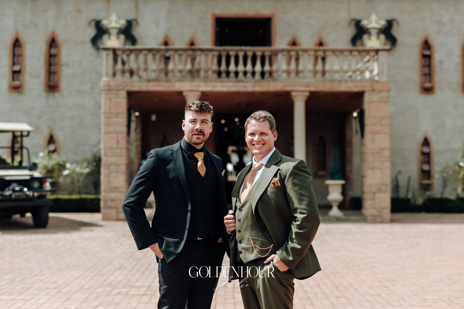 Two groomsmen pose confidently outside elegant venue, one in black suit, one in olive green suit.