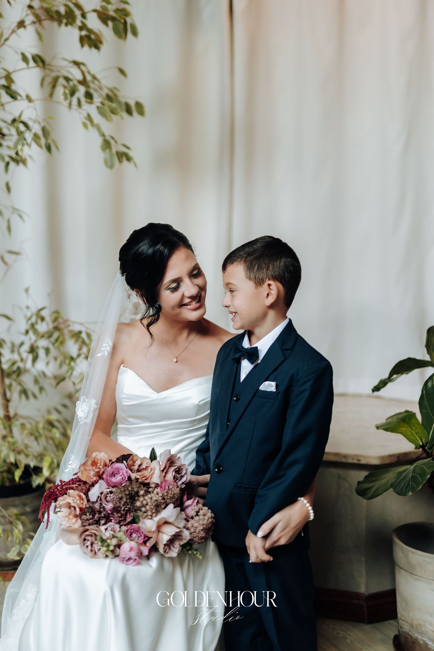 Bride in strapless white gown sharing tender moment with young boy in navy suit holding floral bouquet.
